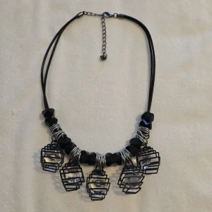 Svart och silver halsband med burdetaljer - Unikt halsband med svarta och silverfärgade detaljer. Halsbandet har fem burformade hängen med klara stenar inuti, vilket ger en modern och edgy look. Perfekt för att ge en extra touch till din outfit.