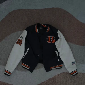 Cincinnati Bengals svart och vit Varsity Jacka - Säljer en cool svart och vit bomberjacka med Cincinnati Bengals-logga. Jackan har långa ärmar och knappar framtill. Den har orange och vita detaljer samt en stor Bengals-logga på ryggen. Perfekt för sportfantasten!