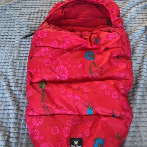 Röd mönstrad åkpåse från Elodie Details - Säljer en färgglad och mysig röd åkpåse från Elodie Details med blommigt och fågelmönster. Perfekt för att hålla ditt barn varmt under kalla dagar. Åkpåsen har en dragkedja och justerbar huva för extra komfort.