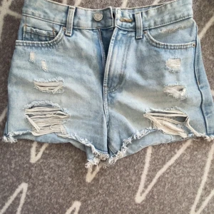 Ljusa jeansshorts med slitningar - Snygga ljusblå jeansshorts med coola slitningar och fransiga kanter. De har en klassisk femficksdesign och stängs med dragkedja och knapp. Perfekta för en avslappnad stil.