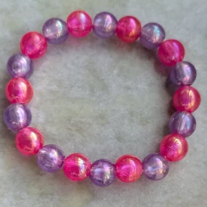 Färgglatt pärlarmband - Säljer ett färgglatt armband med skimrande pärlor i rosa och lila. Perfekt för att lägga till en pop av färg till din outfit. Armbandet är elastiskt och passar de flesta handleder. Passar barn 