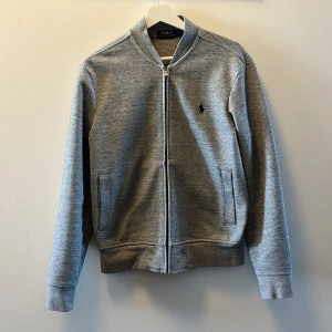 Grå zip-tröja från Polo Ralph Lauren - Säljer en stilren grå zip-tröja från Polo Ralph Lauren. Tröjan har en dragkedja framtill och en liten broderad logotyp på bröstet. Perfekt för en avslappnad och snygg look.