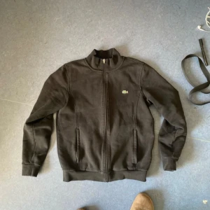 Svart jacka från Lacoste Sport - Säljer en svart jacka från Lacoste Sport med dragkedja och ribbade muddar. Jackan har en liten broderad logotyp på bröstet och en hög krage. Perfekt för en sportig look!