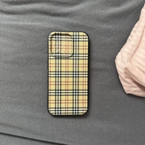 Burberry skal - Snyggt mobilskal med klassiskt rutigt mönster i beige, svart och rött. Skalet har en slimmad design och skyddar baksidan av din telefon. Perfekt för att ge din mobil en stilren look. iPhone 15 PRO