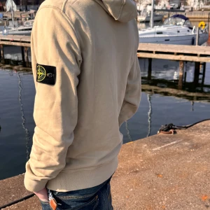 Beige hoodie från Stone Island - Säljer en snygg beige hoodie från Stone Island med deras ikoniska märke på ärmen. Tröjan har en känguruficka och justerbar huva med dragsko. Perfekt för en avslappnad stil vid vattnet eller i stan.