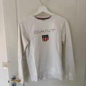 Vit långärmad tröja från GANT - Säljer en vit långärmad tröja från GANT med logotyp och emblem på bröstet. Tröjan har en rund halsringning med röd och blå randig detalj på insidan. Storlek: barn 158/164.