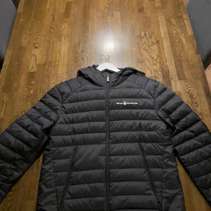 Svart dunjacka från Sail Racing - Säljer en svart dunjacka från Sail Racing med huva och dragkedja. Jackan har en stilren design med Sail Racing-logga på bröstet och baksidan. Perfekt för kyligare dagar. Använd ett par gånger men inte alls så ofta, fräsch och väl omhändertagen!