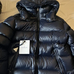 Mörkblå dunjacka från Moncler - Säljer en snygg mörkblå dunjacka från Moncler. Jackan har en glansig finish och är utrustad med dragkedja och huva. Perfekt för kyliga dagar med sin pufferdesign som håller dig varm och stilren.