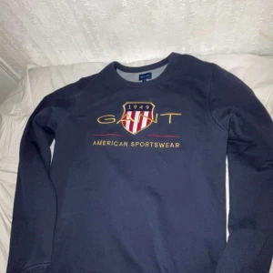 Mörkblå sweatshirt från GANT - Säljer en snygg mörkblå sweatshirt från GANT med ett broderat emblem och texten 'American Sportswear' på framsidan. Tröjan har långa ärmar och en rund halsringning. Perfekt för en avslappnad stil.