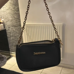 Svart axelväska från Juicy Couture - Snygg svart axelväska från Juicy Couture med guldfärgade detaljer och kedja. Väskan har en dragkedja upptill och är perfekt för att bära dina nödvändigheter med stil. Den kompakta designen gör den lätt att bära med sig överallt.
