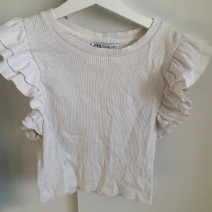 Vit ribbad topp från Zara - Säljer en söt vit ribbad topp från Zara med volangdetaljer på ärmarna. 