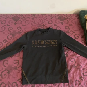Svart tröja från Hugo Boss - Säljer en stilren svart tröja från Hugo Boss med guldglittrig text på bröstet. Tröjan har långa ärmar och en rund halsringning. Perfekt för en elegant och avslappnad look. Storlek S