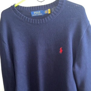 Marinblå tröja från Polo Ralph Lauren - Säljer en stilren marinblå tröja från Polo Ralph Lauren med det ikoniska röda logotypbroderiet på bröstet. Tröjan har en rund halsringning och är tillverkad i ett mjukt material som ger en bekväm passform. Perfekt för en klassisk look.