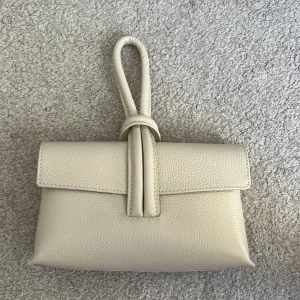 Beige handväska med knyt - Säljer en stilren beige handväska med unik knytning på handtaget. Justerande axelband tillkommer. Nyskick! Ordenarie pris 999kr.