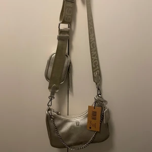 Silver axelväska med kedjedetalj Steve Maden - Snygg silverfärgad axelväska med justerbar axelrem och kedjedetalj. Väskan har en dragkedja och en liten extra ficka på remmen. Perfekt för att bära med sig det viktigaste med stil.