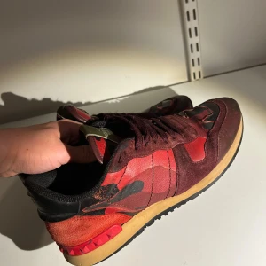Röda valentino rockrunners  - Snygga röda sneakers från Valentino med ett coolt camouflage-mönster. Skorna har en kombination av mocka och syntetmaterial, med svarta och röda detaljer. Sulan är i en kontrasterande beige färg och har ett greppvänligt mönster. Hör av dig vid minsta fundering!