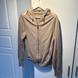 Beige sip-hoodie från Burberry - Säljer en snygg beige, cashmere hoodie från Burberry med dragkedja och huva. Tröjan har långa ärmar och ett diskret broderat märke på framsidan. Fodret i fickorna har ett klassiskt rutigt mönster. Perfekt för en stilren och avslappnad look. Skriv för fler bilder/om du har några frågor. (Säljs i befintligt skick)
