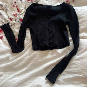 Svart ribbad kofta - Säljer en svart ribbad kofta med knappar framtill. Den är croppad och har långa ärmar. Perfekt för lager-på-lager-stil eller som ett lättare plagg för svalare dagar.