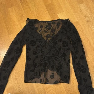 Svart blommig blus - Säljer en elegant svart blus med blommigt mönster. Blusen är transparent och har långa ärmar samt en knytning framtill. Perfekt för en stilfull look. Använd max 2 gånger!!