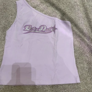 Ljuslila oneshoulder topp med tryck - Säljer en ljuslila oneshoulder topp från New Look med ett glittrigt 'Baby Doll' tryck och stjärndetaljer. Perfekt för en unik och trendig stil. Tillverkad i Turkiet.