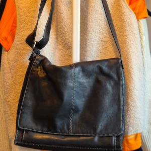 Svart skinnväska med axelrem - Snygg svart axelväska i äkta skinn med flera praktiska fack och dragkedjor. Väskan har en justerbar axelrem och stilren design med italiensk stil. Perfekt för att bära med sig det nödvändigaste med stil.