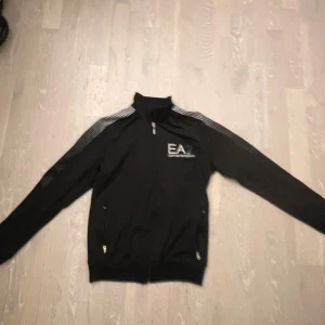Svart jacka från Emporio Armani - Säljer en stilren svart jacka från Emporio Armani med dragkedja och vita detaljer. Jackan har långa ärmar och en hög krage. Perfekt för en sportig look eller avslappnade dagar.