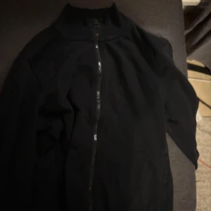 Svart tröja  från BOSS - Säljer en stilren svart jacka från BOSS med dragkedja framtill. Jackan har en hög krage och långa ärmar, perfekt för en minimalistisk look. Passar bra till både vardag och mer uppklädda tillfällen.