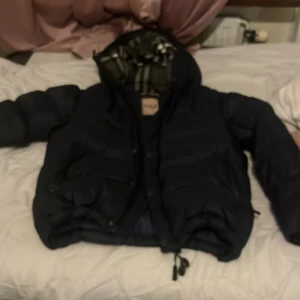 Burberry - Säljer en snygg mörkblå dunjacka från Burberry med klassiskt rutigt foder. Jackan har en huva och praktiska fickor framtill. Perfekt för kyliga dagar.
