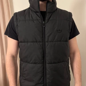 Svart dunväst från Adidas - Snygg svart dunväst från Adidas med dragkedja och diskret logga på bröstet. Västen har en hög krage och klassiska vita ränder längs sidorna. 
