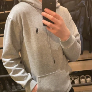 Grå hoodie från Polo Ralph Lauren - Säljer en stilren grå hoodie från Polo Ralph Lauren med klassisk känguruficka och justerbar huva. Tröjan har ribbade muddar och en liten broderad logga på bröstet. Perfekt för en avslappnad look.