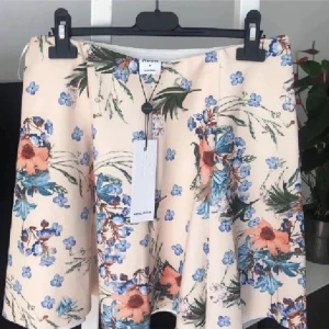 Blommig kjol M - Söt blommig kjoli en ljus beige nyans med blå och orange blommor. Kjolen har en kort längd och en lätt utsvängd form som ger en fin siluett. Perfekt för varma dagar!