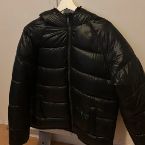 Svart pufferjacka från Demand - Säljer en svart pufferjacka från Demand med en cool patch på ärmen. Jackan har en dragkedja framtill och är perfekt för kyligare dagar. Den är långärmad och har en modern look.