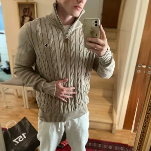 Beige stickad tröja med dragkedja från Morris - Säljer en beige stickad tröja från Morris med kabelstickat mönster och halv dragkedja framtill. Tröjan har hög krage och ribbade muddar. Perfekt för lager-på-lager och snygg detalj med Morris-loggan på bröstet.