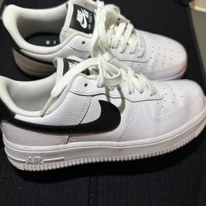 Nike Air Force 1 sneakers i vitt och svart - Säljer ett par klassiska Nike Air Force 1 sneakers i vitt skinn med svart swoosh. Skorna har snörning, perforerad tå och platt sula. Perfekta för dig som gillar stilrena och tidlösa sneakers. Använda 2-3 gånger bara!