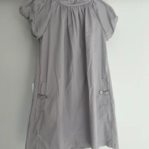 Ljusgrå klänning från By Malene Birger - Säljer en ljusgrå klänning från By Malene Birger med korta puffärmar och två fickor framtill med knappdetaljer. Klänningen har en lös passform och är tillverkad i 100% bomull, perfekt för en stilren och avslappnad look.