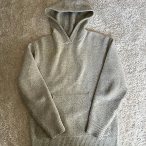 stickad hoodie - Mysig ljusgrå stickad hoodie med huva och magficka. Tröjan har långa ärmar och ribbade muddar vid ärmslut och nederkant. Perfekt för kyliga dagar när du vill vara både bekväm och snygg. Storlek S