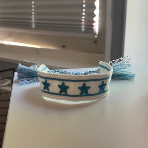 Vitt och blått armband med stjärnor - Ett handgjort armband i vitt tyg med blå broderade stjärnor och blå detaljer. Armbandet har knytning och tofsar i ändarna för justerbar passform. Perfekt accessoar för att addera lite färg till din stil.
