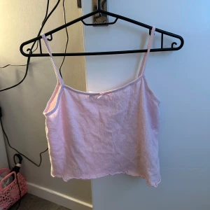 Ljusrosa ribbad linnetopp - Söt ljusrosa linnetopp med smala axelband och ribbad struktur. Toppen är croppad och har en liten rosett framtill. Perfekt för varma dagar eller som ett lager under andra plagg. Nattlinne