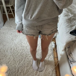 Grå slitna jeansshorts med fransar - Grå jeansshorts med råa kanter och slitningar, klassisk femficksmodell och hög midja. Shortsen har fransar längs bensluten och flera slitna detaljer framtill och baktill för en tuff look.