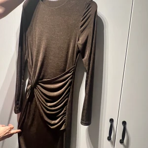 Brun sammetsklänning med rynkade detaljer - Säljer en brun långärmad klänning i mjuk sammet med rynkade detaljer på ena sidan. Klänningen har en kroppsnära passform och rund halsringning. Perfekt för dig som vill ha en stilren men ändå unik look.