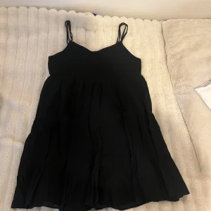 Svart sommarklänning med smock och tunna axelband - Säljer en svart klänning med smock upptill och tunna axelband. Klänningen har en luftig passform och är perfekt för varma dagar. Den är kort i modellen och har en enkel, stilren design.