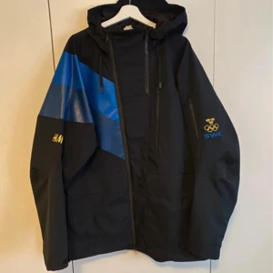 Svart och blå vindjacka från H&M x OS Sverige - Säljer en svart vindjacka med blå detaljer från H&M:s officiella OS-kollektion för Sverige. Jackan har huva, dragkedja framtill, bröstfickor med dragkedja och tryck med OS-ringarna och 'SWE' på ärmen samt 'SVERIGE' i guld på ryggen.