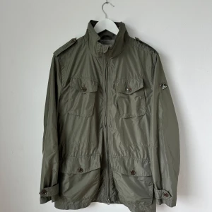 Olivgrön Foreman vindjacka från J.Lindeberg - Säljer en olivgrön vindjacka från J.Lindeberg med fyra fickor framtill, axelklaffar och hög krage. Jackan har dragkedja och knappar samt är helfodrad i grått. Perfekt för vår och höst. Modell: Foreman 53 2L Tech Poly - Artnr: X235