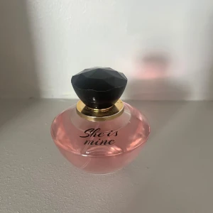 La Rive: She is mine parfym - En söt parfymflaska med rund form och rosa glas. Flaskan har en svart, diamantformad kork och en guldfärgad ring vid halsen. Texten 'She is mine' står på framsidan. Perfekt för dig som gillar feminina och fräscha dofter.