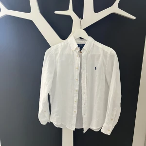 Vit linneskjorta från Polo Ralph Lauren strl M (junior) - Klassisk vit skjorta i linne från Polo Ralph Lauren med button-down krage och broderad logga på bröstet. Skjortan har långa ärmar och knäppning framtill. Perfekt för en stilren och fräsch look. Strl M junior (10-12 år). 