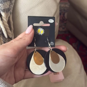 Svart och guldiga statement örhängen - Stora, runda örhängen med svart bas och droppformade detaljer i guld och beige. Perfekta för att ge en cool touch till din outfit. Hängande modell med metallkrok. Nya!