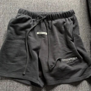 Svarta shorts från Essentials Fear of God - Svarta shorts från Essentials Fear of God med elastisk midja och snörning. Mjukt material och logga tryckt på vänster ben samt en liten patch framtill. Perfekta för en avslappnad stil.