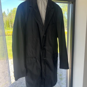 Svart blazer/jacka från Oscar Jacobson - Klassisk svart coat från Oscar Jacobson med tre knappar framtill och innerfoder i ljusgrått med tunna ränder. Kavajen har fickor med lock och en bröstficka. Perfekt för formella tillfällen. Otroligt stilren med slimmade armslut. Storlek 50/M