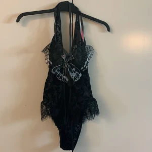 Svart spetsig body med fjärilsdetalj - Säljer en svart body i spets med en stor broderad fjäril framtill och matchande ärmar med tyll. Bodyn har djup urringning och fina spetsdetaljer, perfekt för att sticka ut. Säljs tillsammans med avtagbara ärmar i samma material. rekommenderar att du syr in tyg eller har top utan armar under i svart