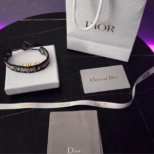Blå armband från Dior - Säljer ett svart armband från Dior med vit broderad logga. Justerbar knytning för perfekt passform. Kommer med originalförpackning, påse och band med Dior-tryck. Perfekt accessoar för att lyfta din stil.